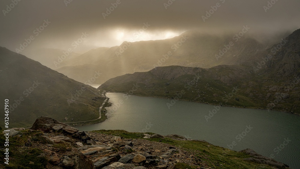 Aerial shot of light shining through gray clouds over a lake, surrounded by rocky mountains