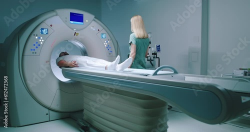 Male patient lies at TC scanner bed. Man goes out of MRI capsule. Female doctor dressed up in uniform have talk with patient. Medical worker smiling and pushes buttons of MRI capsule. photo