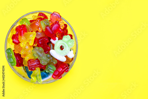 A large pile of multi-colored jelly gummy candies in a glass bowl on a yellow background. Variety of delicious gummies. Various tasty sticky candies in a glass bowl close-up. Free space for text
