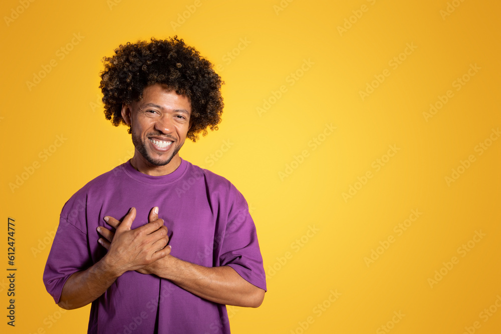 Cheerful black adult curly man in purple t-shirt makes gratitude gesture, put hands to chest Cheerful black adult curly man in purple t-shirt makes gratitude gesture, put hands to chest