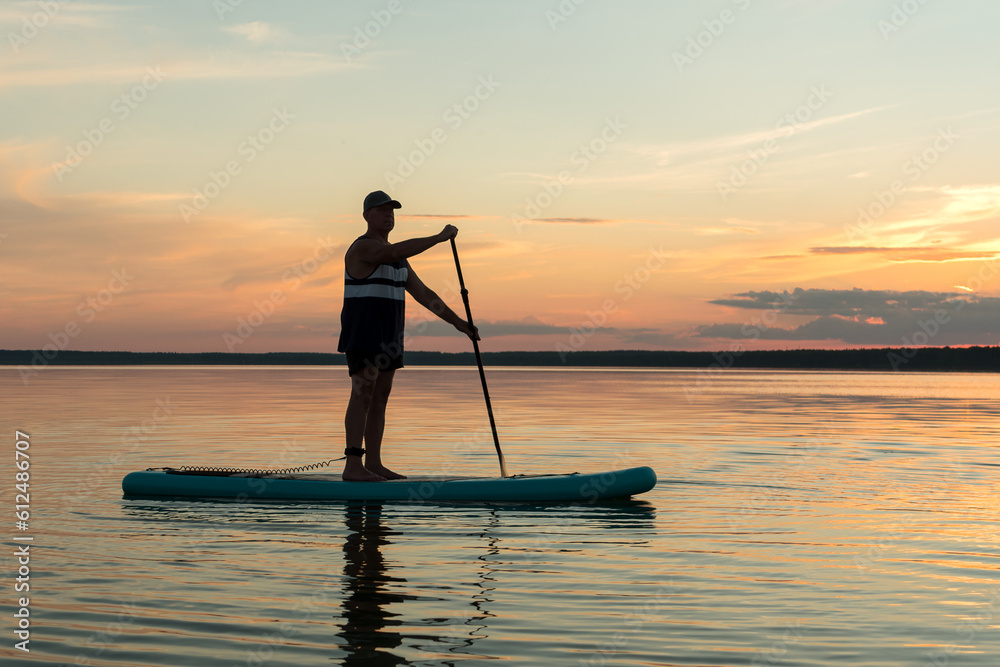 A man in shorts on a SUP board with a paddle at sunset swims in the water of the lake against the background of a purple pre-sunset sky.
