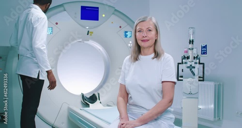 Shooting with smooth zoom. Mature woman sits at TC scanner bed. African American doctor adjusts MRI capsule behind woman. Woman dressed up in white is looking at camera. photo