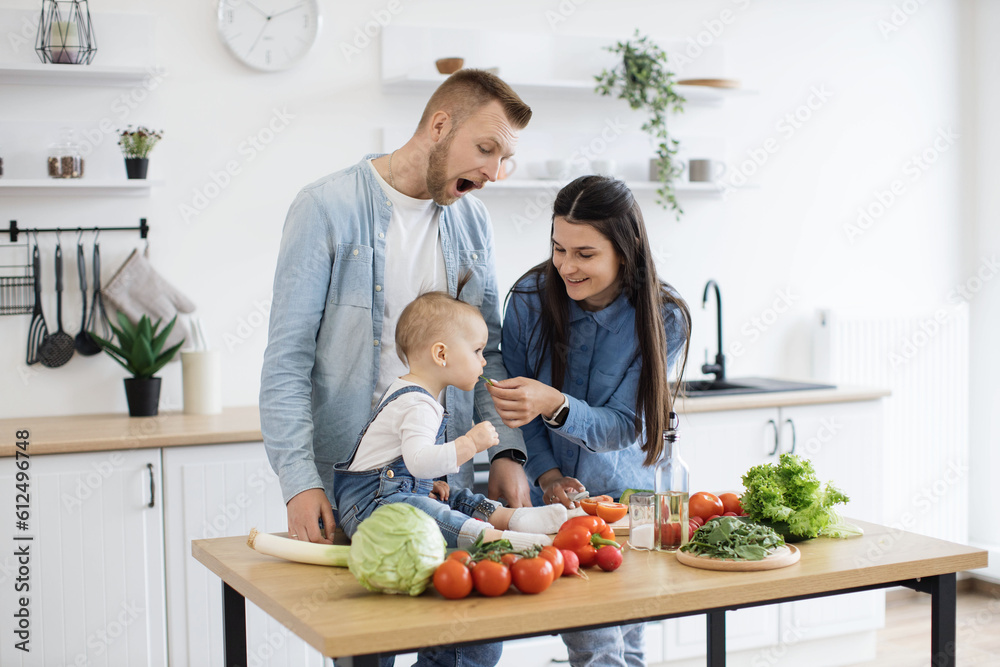 Caring young woman in denim clothes letting baby girl taste green vegetable while cooking dishes for dinner at noon. Mindful parents getting child used to new textures and new flavours of solid food. Caring young woman in denim clothes letting baby girl taste green vegetable while cooking dishes for dinner at noon. Mindful parents getting child used to new textures and new flavours of solid food.
