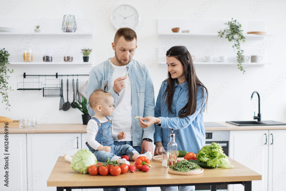 Beautiful fit woman offering slice of peeled cucumber to little kid while curious daddy looking shocked standing aside. Attentive mother giving daughter new taste of solid food while preparing salad. Beautiful fit woman offering slice of peeled cucumber to little kid while curious daddy looking shocked standing aside. Attentive mother giving daughter new taste of solid food while preparing salad.