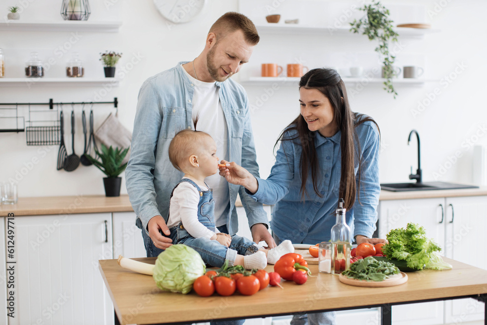 Happy caucasian couple in denim clothes feeding small child with slice of red tomato during lunch time at home. Curious baby girl checking benefits of vegetable rich in vitamin C for proper nutrition. Happy caucasian couple in denim clothes feeding small child with slice of red tomato during lunch time at home. Curious baby girl checking benefits of vegetable rich in vitamin C for proper nutrition.