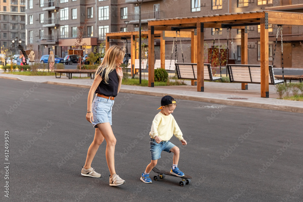 Happy family enjoying spending time together skateboarding at summer on a city street at sunset. Family activities concept. Mother and little son riding with skateboard.