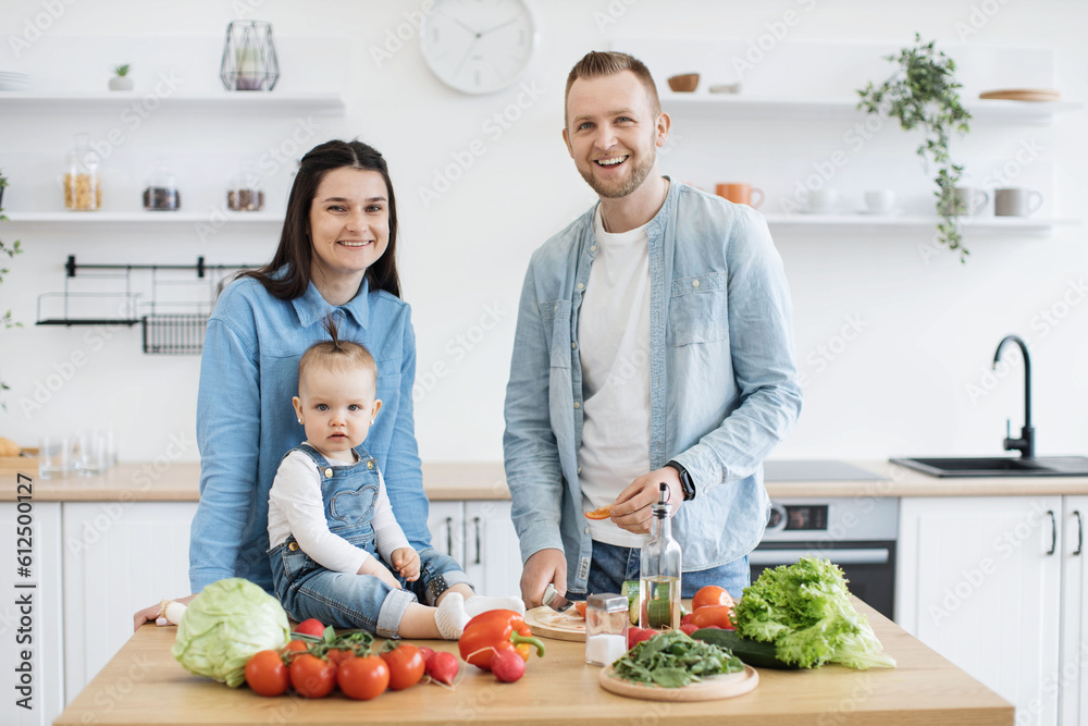 Portrait of happy three-person family in denim wear standing at kitchen table with assorted vegetables and greens. Stylish spouses achieving balanced diet by using organic products for home cooking. Portrait of happy three-person family in denim wear standing at kitchen table with assorted vegetables and greens. Stylish spouses achieving balanced diet by using organic products for home cooking.