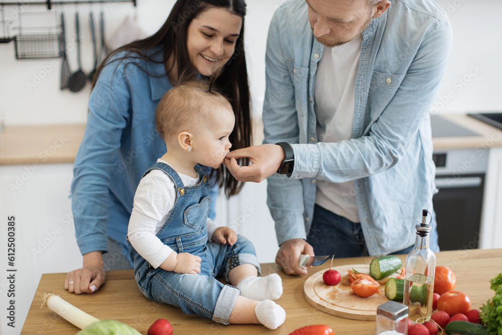 Close up view of beautiful little girl in cozy outfit eating tasty slice held by caring father while mom smiling behind. Young couple weaning baby girl by giving solid food while making vegan meals. Close up view of beautiful little girl in cozy outfit eating tasty slice held by caring father while mom smiling behind. Young couple weaning baby girl by giving solid food while making vegan meals.