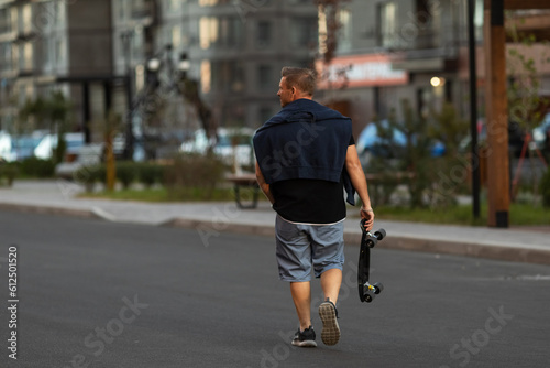 Back view young man walking down the street with skateboard in his hands. Close up portrait of a young male carrying his longboard down the street. © Irina Mikhailichenko