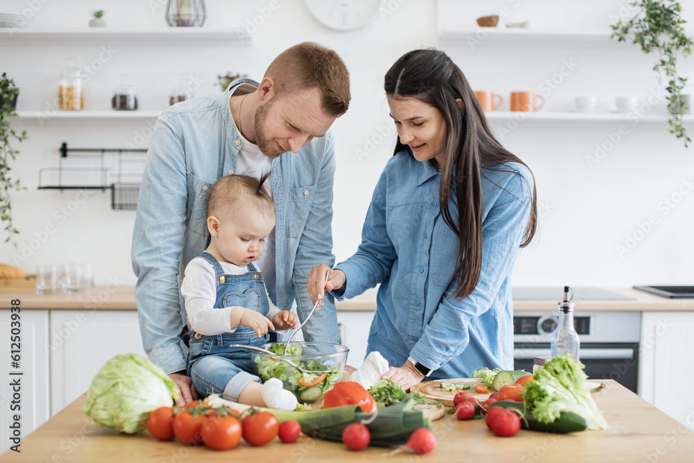 Front view of smiling spouses dressing garden salad while curious little daughter resting in kitchen of apartment. Young caucasian couple and baby enjoying benefits from cooking together as family.