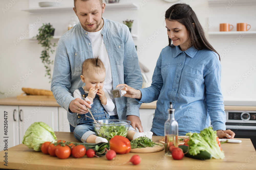 Affectionate young family with little infant having fun together while cooking natural balanced food in kitchen. Happy smiling parents enjoying weekend time with small child. Concept of healthy diet.