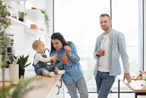 Adorable sweet kid letting mommy to take sip of fresh healthy juice through plastic straw in cozy kitchen. Bearded father holding glass with drink, leaning on table and smiling on camera.