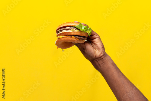 african american male hand holds and shows tasty burger on yellow isolated background, close-up of fast food