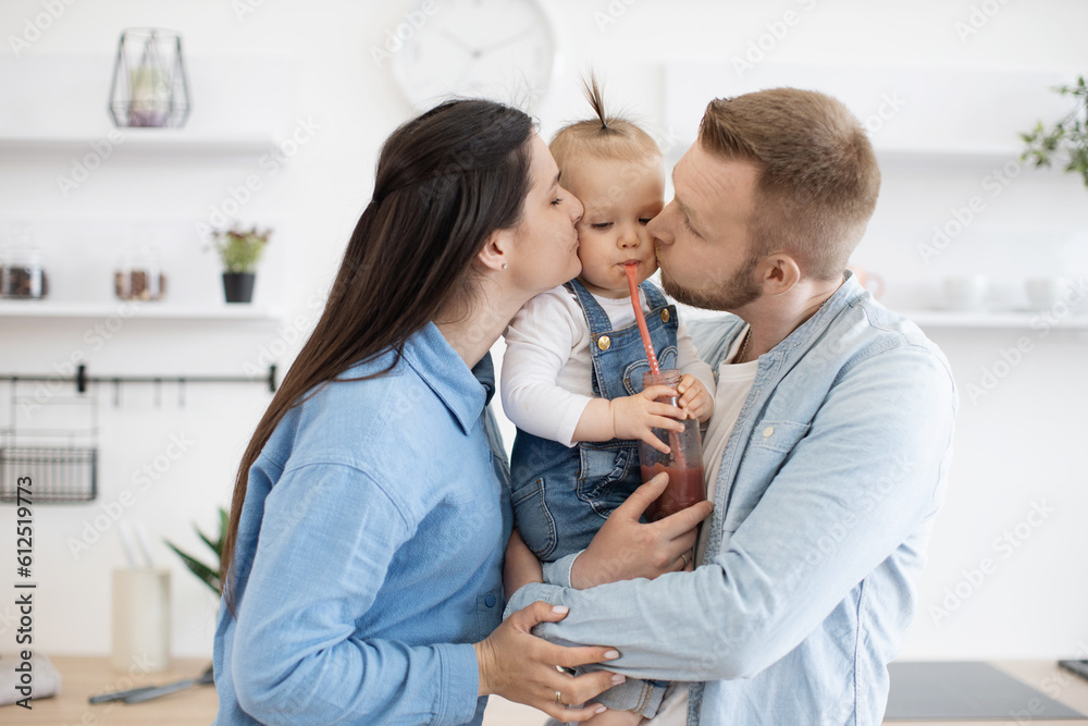 Loving mom and dad in everyday outfit kissing sweet little daughter in cheeks while standing in new modern kitchen. Adorable kid drinking fresh juice through plastic straw. Concept of happy family. Loving mom and dad in everyday outfit kissing sweet little daughter in cheeks while standing in new modern kitchen. Adorable kid drinking fresh juice through plastic straw. Concept of happy family.