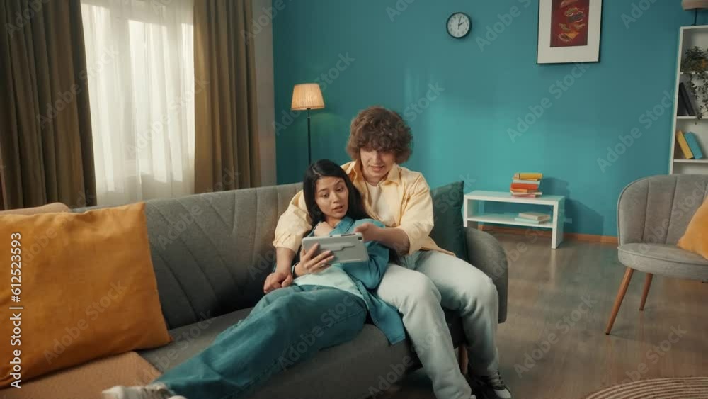 A woman and a man watch a movie, photo, video on a tablet, discuss what they saw. A young Asian woman lies on a sofa with a tablet in her hands, leaning her back on a man. A woman and a man watch a movie, photo, video on a tablet, discuss what they saw. A young Asian woman lies on a sofa with a tablet in her hands, leaning her back on a man.