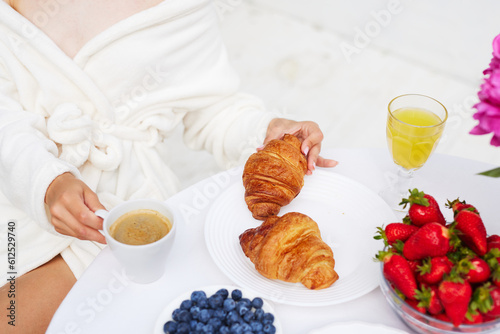 A young woman in a bathrobe, a towel on her head enjoys her vacation at the resort, has breakfast coffee with a croissant and strawberries