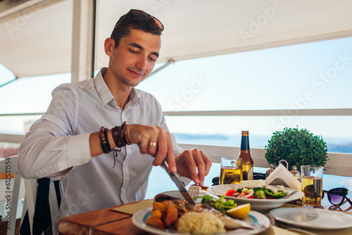Man eats fish with vegetables in outdoors restaurant. Fresh seafood dinner with sea landscape. Summer vacation