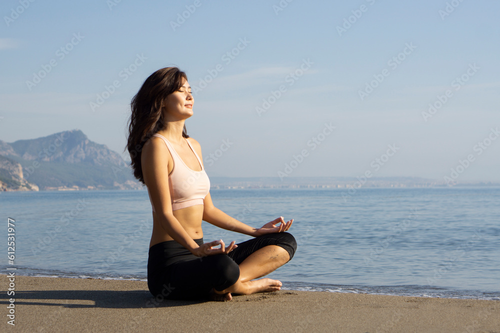 Happy peaceful young asian woman meditating on seashore at sunrise, practicing yoga on sandy beach