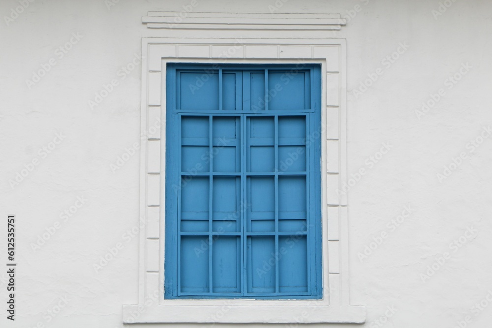 Closeup of the vintage blue window with bars and a white patterned frame on a white concrete wall