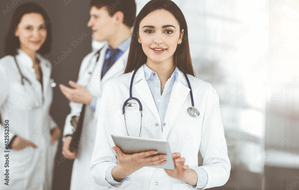 Smiling young woman-doctor is standing in sunny clinic indoors. Smiling young woman-doctor is standing in sunny clinic indoors.