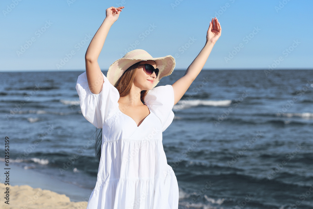 Happy smiling woman in free happiness bliss on ocean beach standing with a hat, sunglasses, and rasing hands. Portrait of a multicultural female model in white summer dress enjoying nature during trav Happy smiling woman in free happiness bliss on ocean beach standing with a hat, sunglasses, and rasing hands. Portrait of a multicultural female model in white summer dress enjoying nature during trav