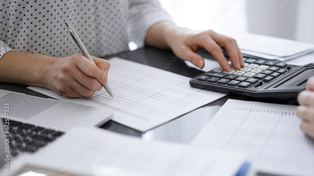 Woman accountant using a calculator and laptop computer while counting taxes with a client or a colleague. Business audit team, finance advisor.