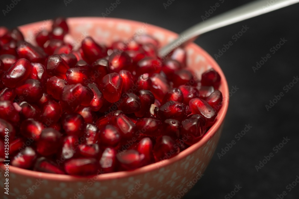 Closeup of a bowl full of red pomegranate juicy seeds, served with a spoon, on the black background