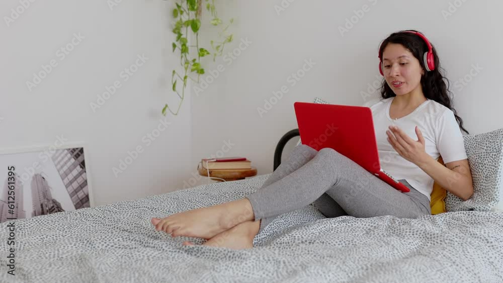 Smiling young Latin American woman with headphones using laptop computer resting on bed in the morning. Millennial girl having a video call on computer. E-learning and education concept. Smiling young Latin American woman with headphones using laptop computer resting on bed in the morning. Millennial girl having a video call on computer. E-learning and education concept.