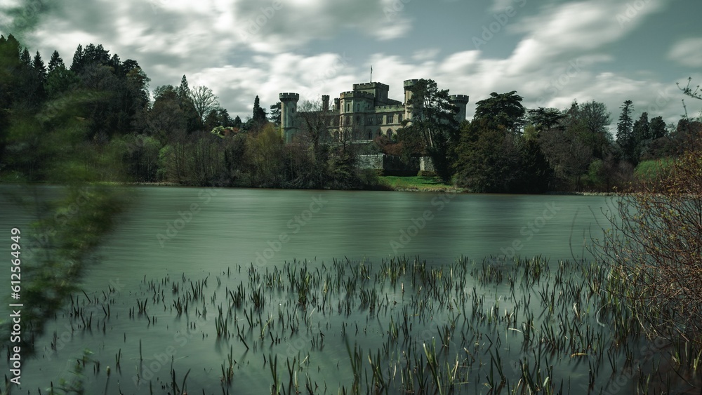 Fototapeta premium Long-exposure waterside view of Eastnor Castle surrounded by trees on a cloudy autumn day