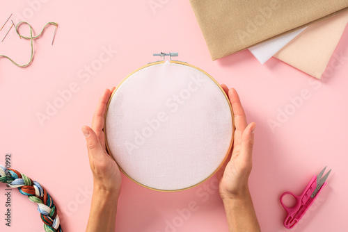 Embroidery concept. Female hands holding round wooden frame with blank canvas over pink background with needlework accessories.