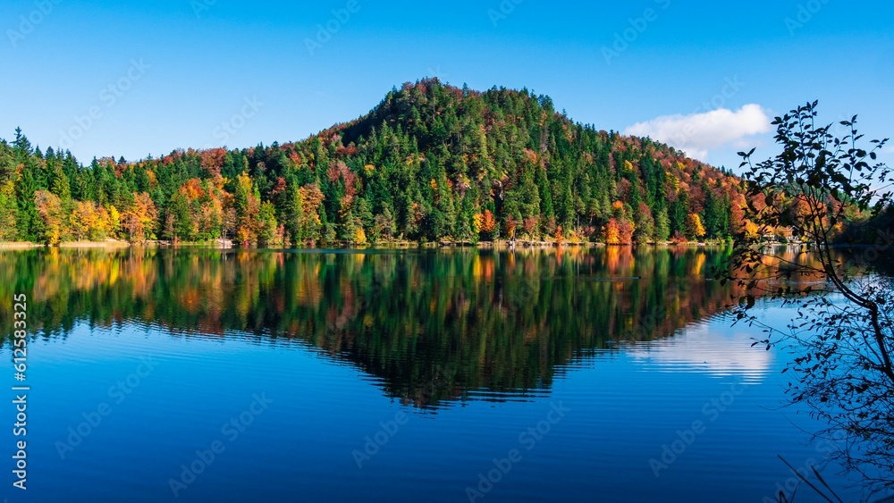 Image of a mount covered by colorful trees with the reflection of it on the lake under the blue sky.