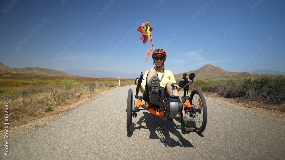 Obraz premium Front view slow motion of elderly senior woman riding recumbent e-bike tricycle bike on a path on a sunny day beside a lake.