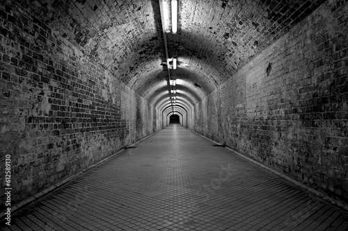 Grayscale of a tunnel with old, dirty walls and lamps on the top of the arched ceilings