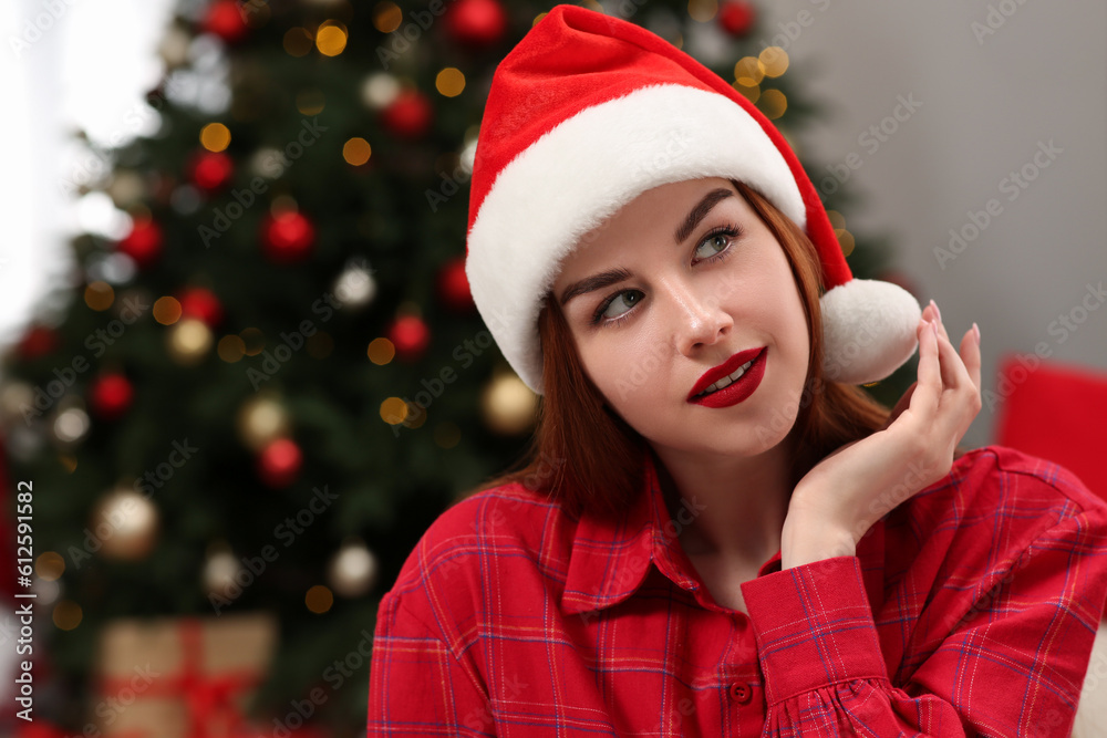 Beautiful young woman in Santa hat near Christmas tree indoors, space for text Beautiful young woman in Santa hat near Christmas tree indoors, space for text