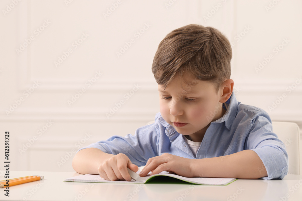 Little boy erasing mistake in his notebook at white desk indoors Little boy erasing mistake in his notebook at white desk indoors