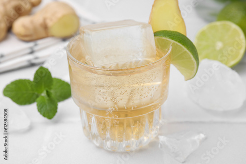 Glass of tasty ginger ale with ice cube and ingredients on white table, closeup