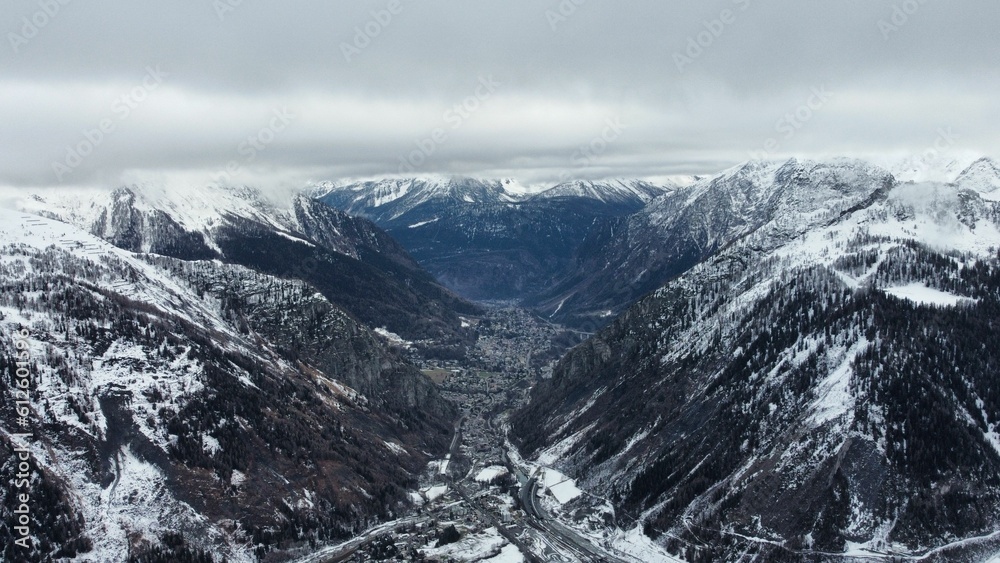 Obraz premium Scenic shot of a village between the snowy mountains covered in evergreen forests on a misty day