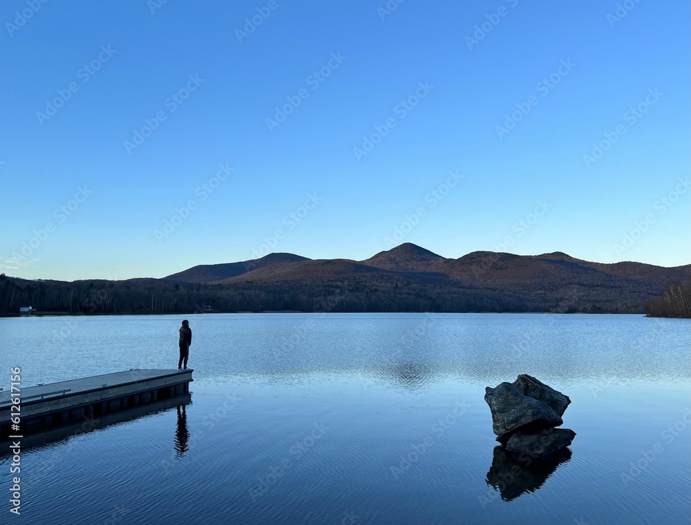 View of a person standing on a wooden pier and a rock in calm water reflecting the clear blue sky