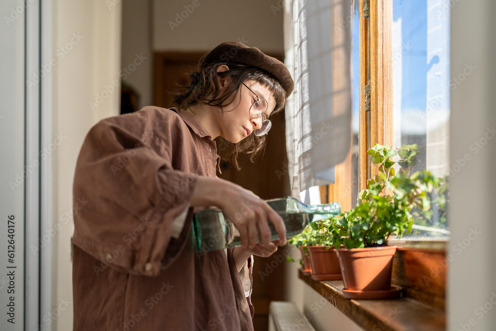 Plant care. Hipster girl watering potted houseplant on windowsill, loving caring for indoor plants, millennial woman holding bottle of water standing by window enjoying indoor gardening hobby Plant care. Hipster girl watering potted houseplant on windowsill, loving caring for indoor plants, millennial woman holding bottle of water standing by window enjoying indoor gardening hobby