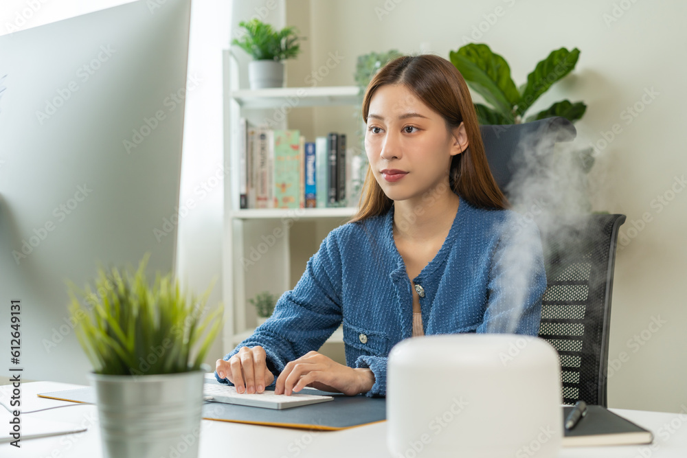 Modern air humidifier device during sitting workplace, happy asian young woman using computer work on internet, enjoying aromatherapy steam scent from essential oil diffuser in room at home office. Modern air humidifier device during sitting workplace, happy asian young woman using computer work on internet, enjoying aromatherapy steam scent from essential oil diffuser in room at home office.