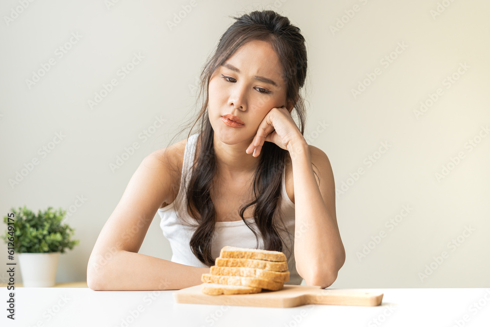 Gluten allergy, asian young woman, girl refusing to eat, looking at bread slice on plate in breakfast food meal on table at home, having a stomach ache. Gluten intolerant and Gluten free diet concept. Gluten allergy, asian young woman, girl refusing to eat, looking at bread slice on plate in breakfast food meal on table at home, having a stomach ache. Gluten intolerant and Gluten free diet concept.