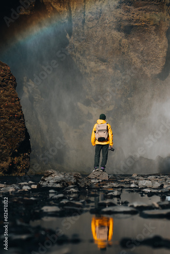 Man in yellow raincoat stands at waterfall as rainbow overhead