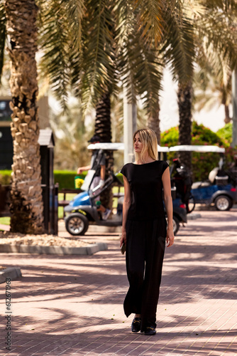 Full length of slim blonde woman in black clothes walking in urban park outdoors at palm trees, looking away. Pretty young lady posing in tropics. Healthy leisure activity concept. Copy ad text space