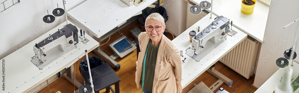Obraz premium Middle aged woman designer looks at camera standing near sewing machines in spacious studio
