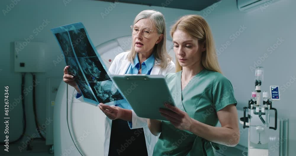 Female doctor and nurse standing at equipped tomography room. Doctor is holding scans and consulting with colleague. Nurse hold folder and listen to doctor carefully. Analizing of examination results,