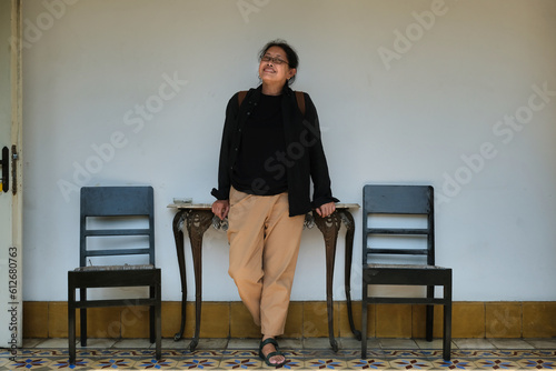 A middle-aged woman in black shirt is standing in front of a table located between two wooden chairs; happy expression photo