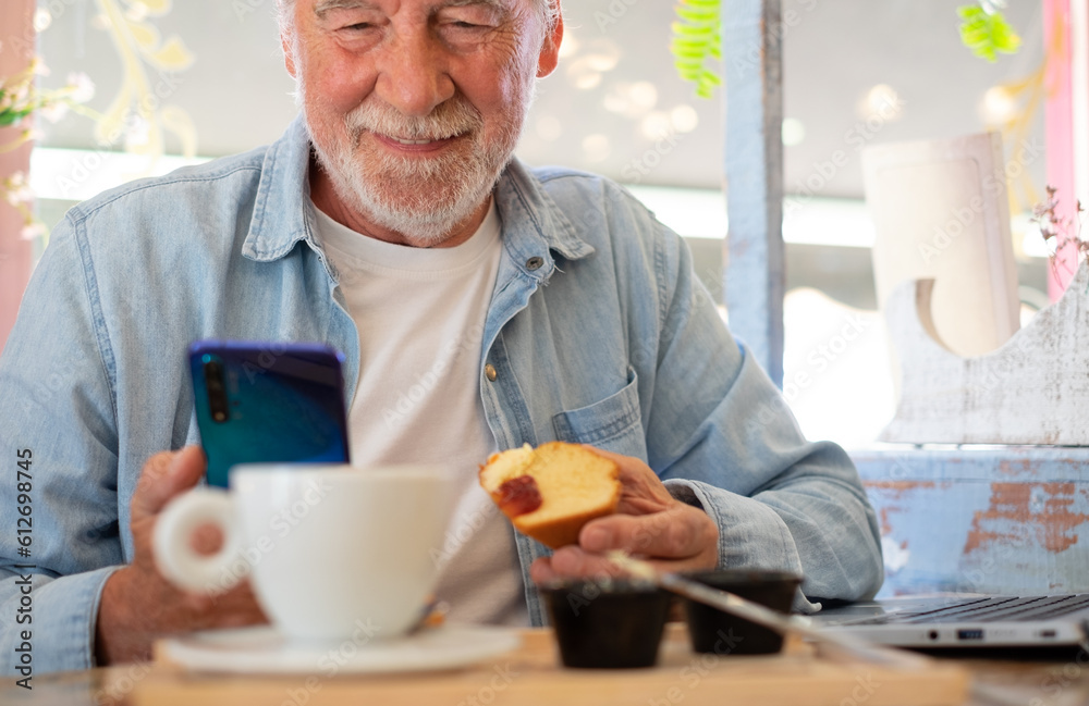 Smiling old senior man in coffee shop having breakfast with cappuccino and sweet food while using phone and laptop. Elderly people enjoying free lifestyle and pleasant moment of relax Smiling old senior man in coffee shop having breakfast with cappuccino and sweet food while using phone and laptop. Elderly people enjoying free lifestyle and pleasant moment of relax