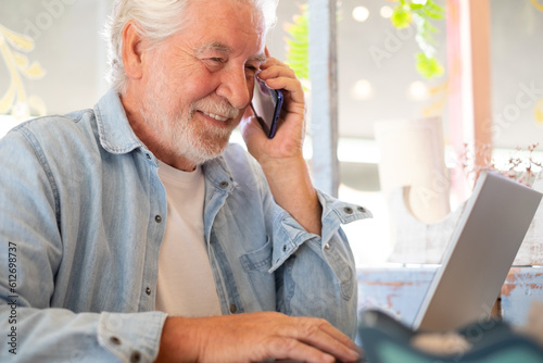 Smart work concept. Smiling senior man in coffee shop while working on laptop talking on phone. Elderly man people enjoying free lifestyle doing job from remote