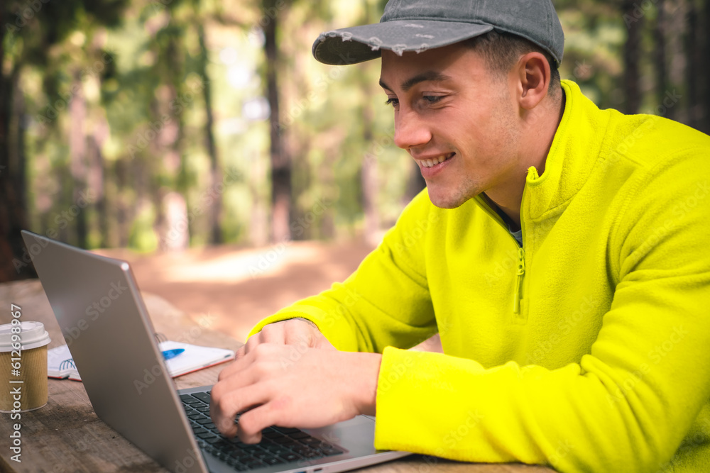Beautiful caucasian boy freelancer working on laptop sitting outdoors in the forest. Hipster young man traveler working distantly while enjoying nature landscape during vacations