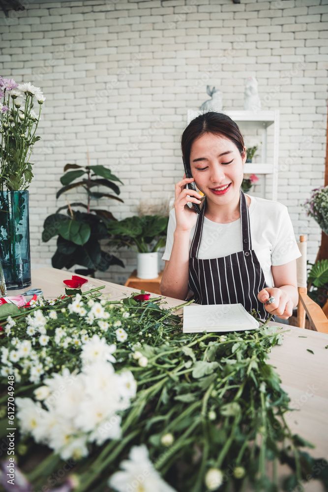 Young florist taking orders on smartphone with smiling face in flower shop full of flowers and supplies. Young florist taking orders on smartphone with smiling face in flower shop full of flowers and supplies.